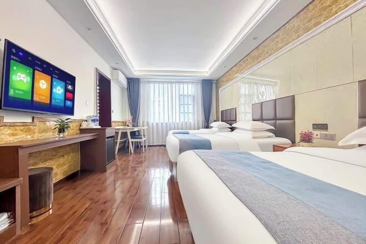 A hotel room with a large flat-screen TV, a desk with a computer, two beds, a small table, and a window with curtains.