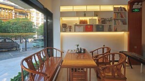 A cozy indoor seating area with a wooden table and chairs, a bookshelf with various items, and a view of the street outside. - No. 1 Inn @ the Corner (Zhangjiajie)