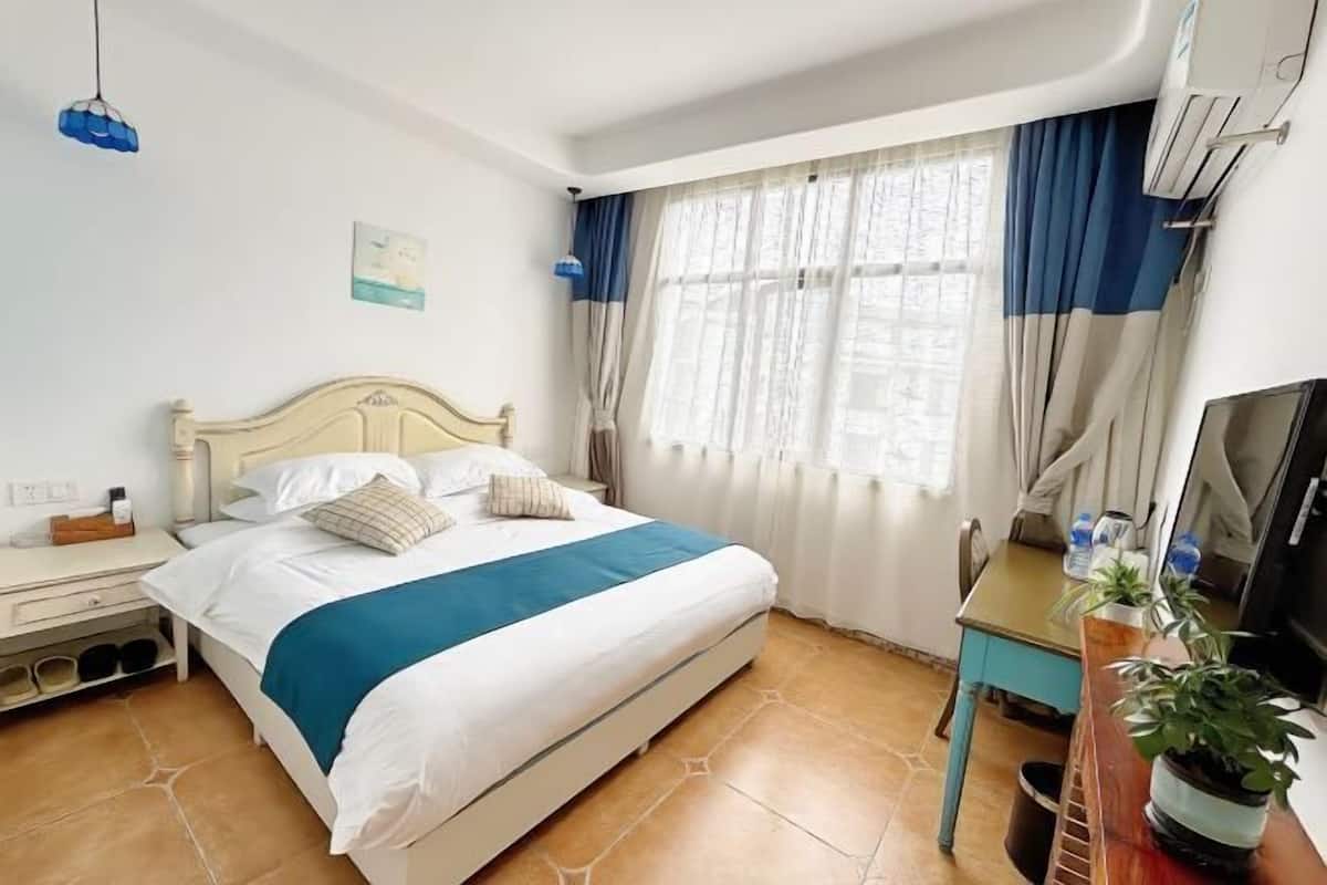A bedroom with a bed, a nightstand, a desk, a chair, a TV, and a window with curtains.