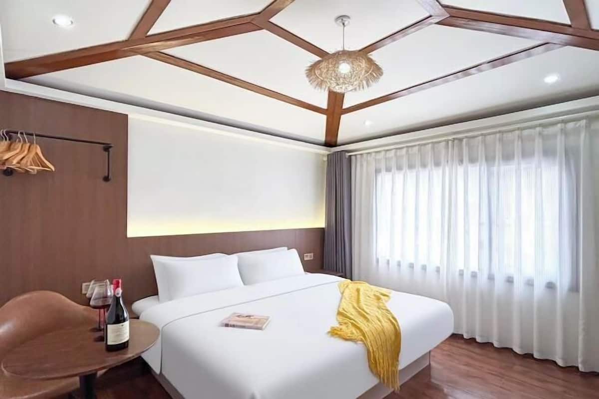 A modern hotel room with a large bed, a wooden ceiling, a hanging light fixture, a window with curtains, and a small table with a bottle of wine.
