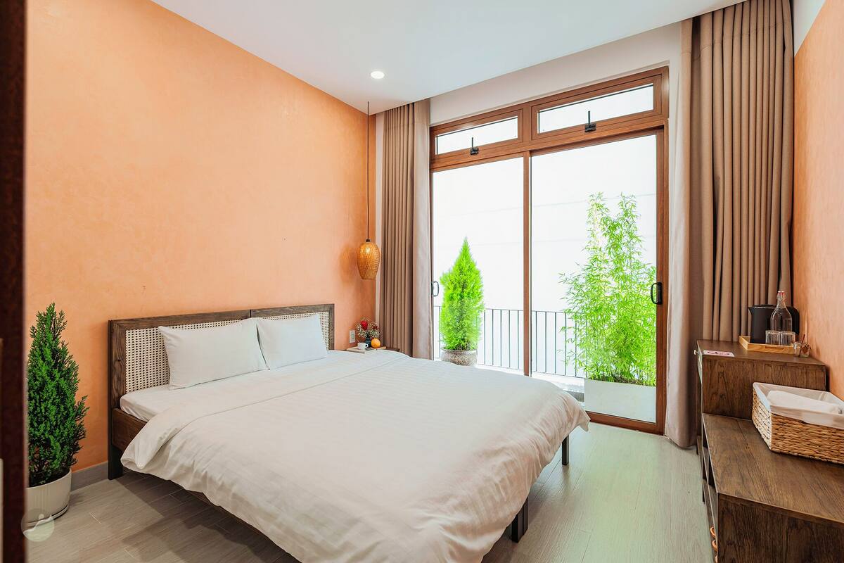 A bedroom with a large bed, a window with curtains, a small table with a bottle, and a potted plant.