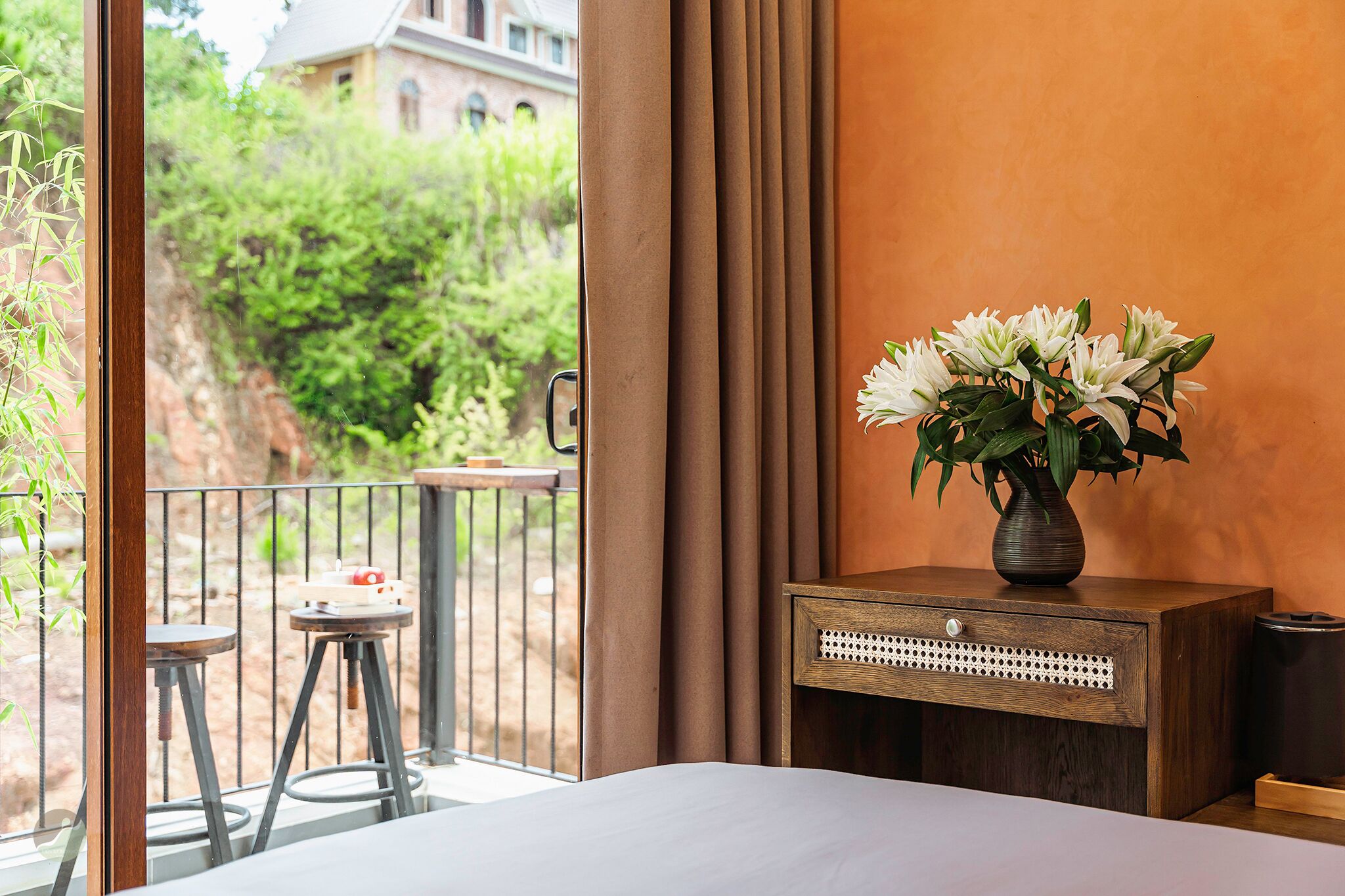 A hotel room with a bed, a small wooden nightstand with a vase of flowers, a balcony with a table and chairs, and a view of greenery and a building outside.