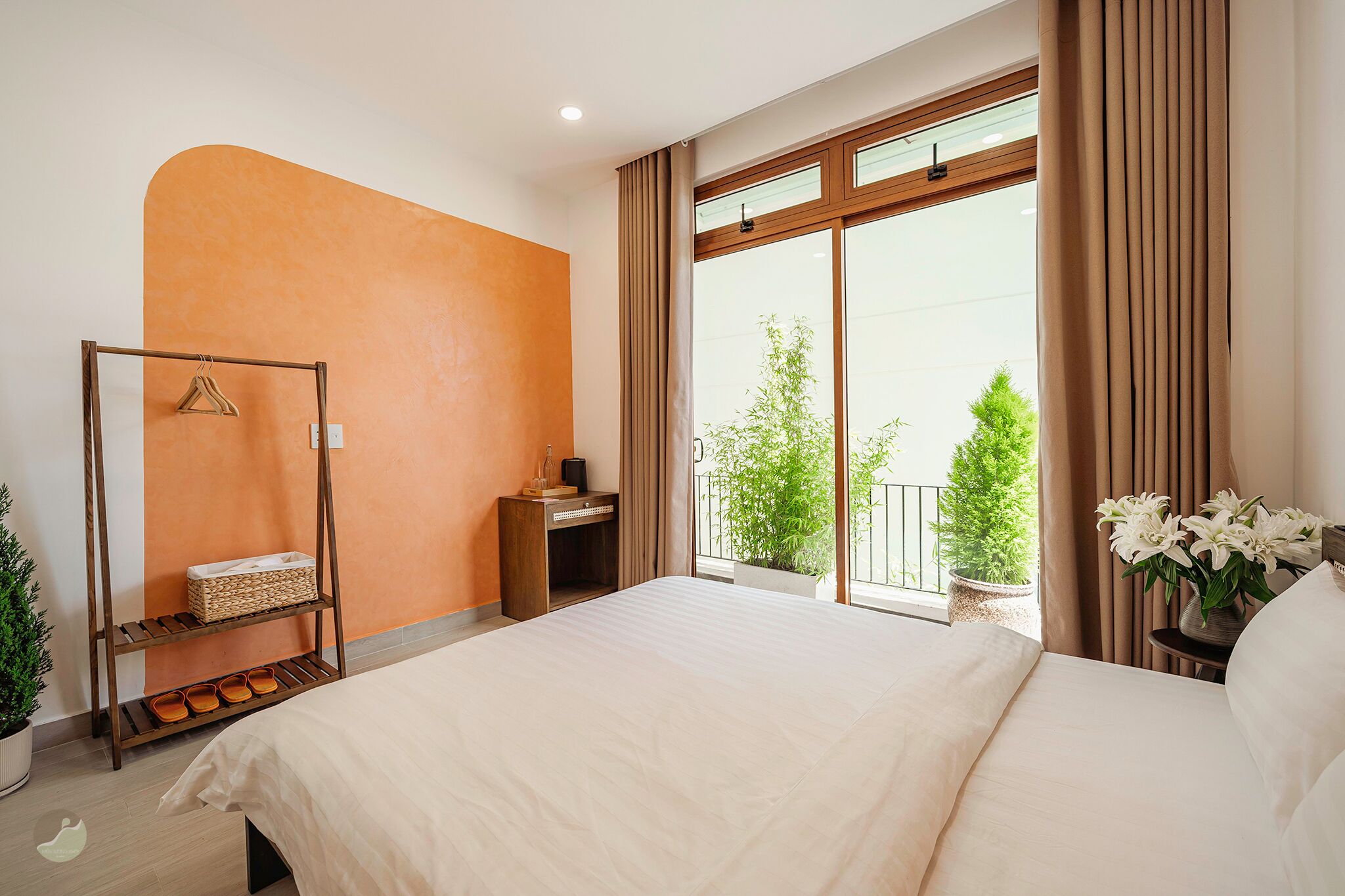 A hotel room with a large bed, a wooden bedside table, a small desk, a hanging shelf, and a view of the outdoors through a sliding glass door.