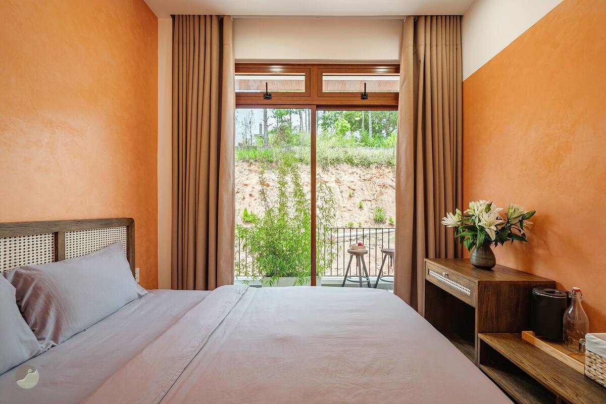 A bedroom with a bed, a bedside table with a vase of flowers, and a view of the outdoors.