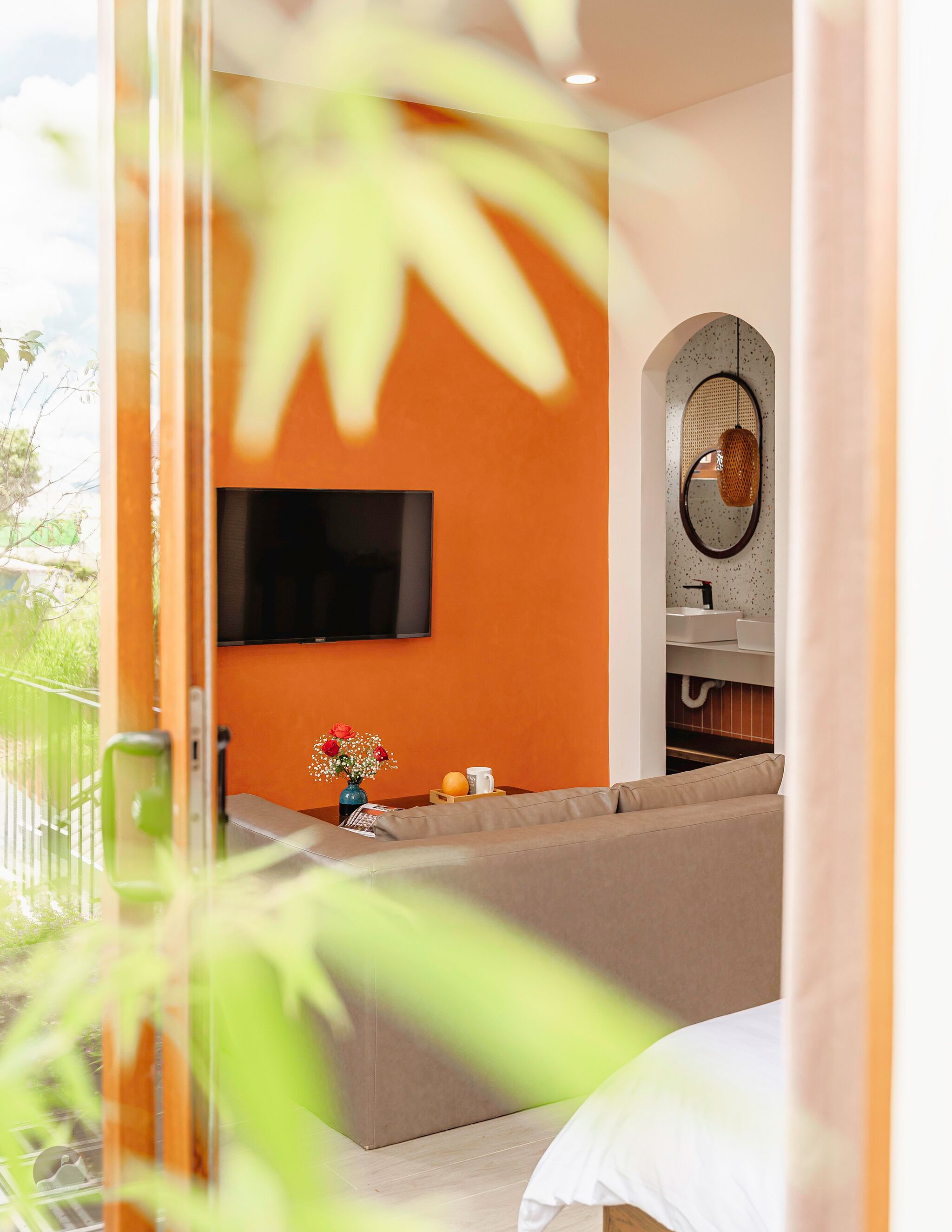 Une chambre moderne avec un grand téléviseur à écran plat fixé sur un mur orange, un canapé, une petite table sur laquelle est posé un vase de fleurs et un luminaire suspendu.