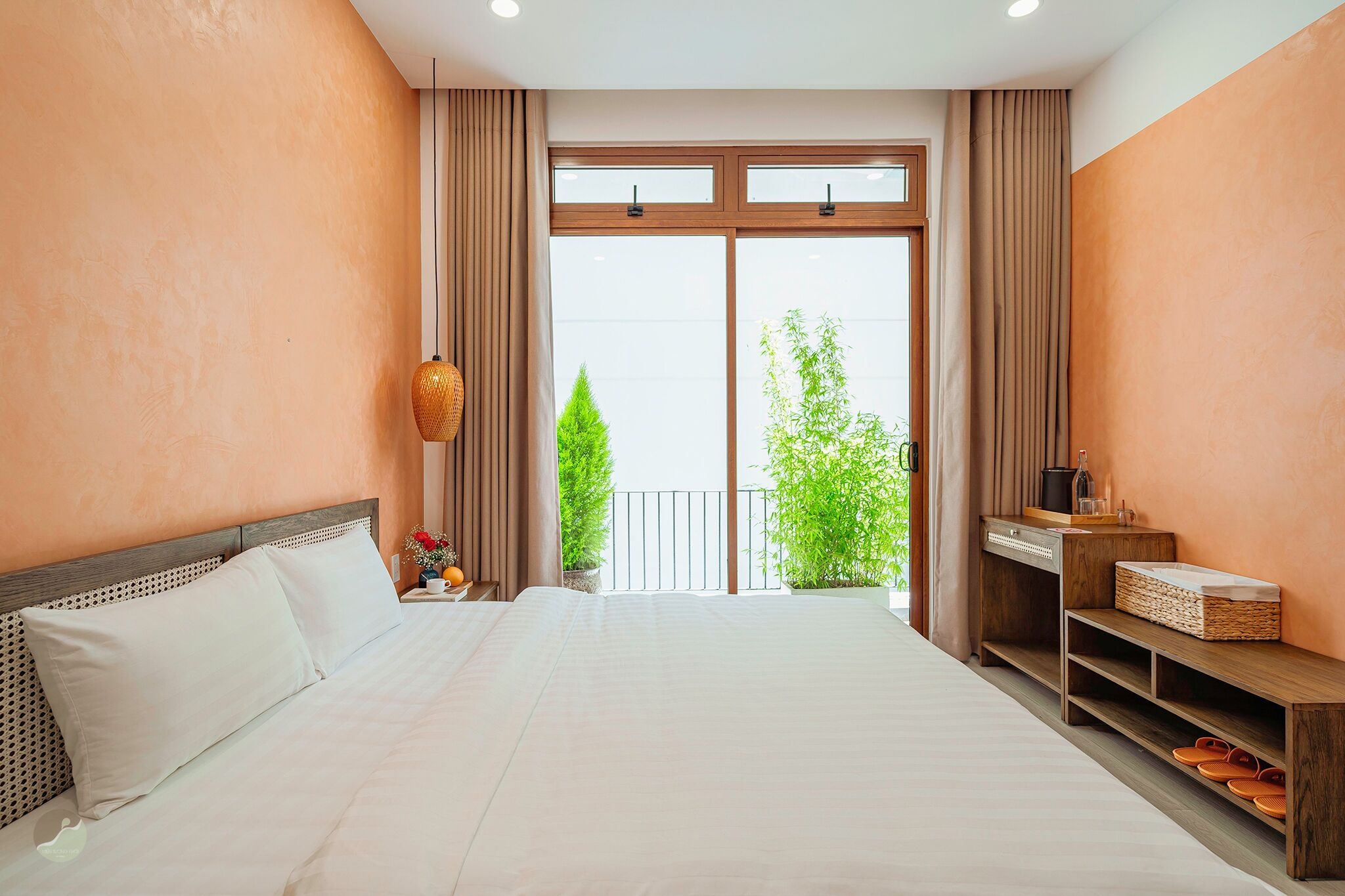 A hotel room with a large bed, a wooden nightstand, a small table, and a view of greenery outside.