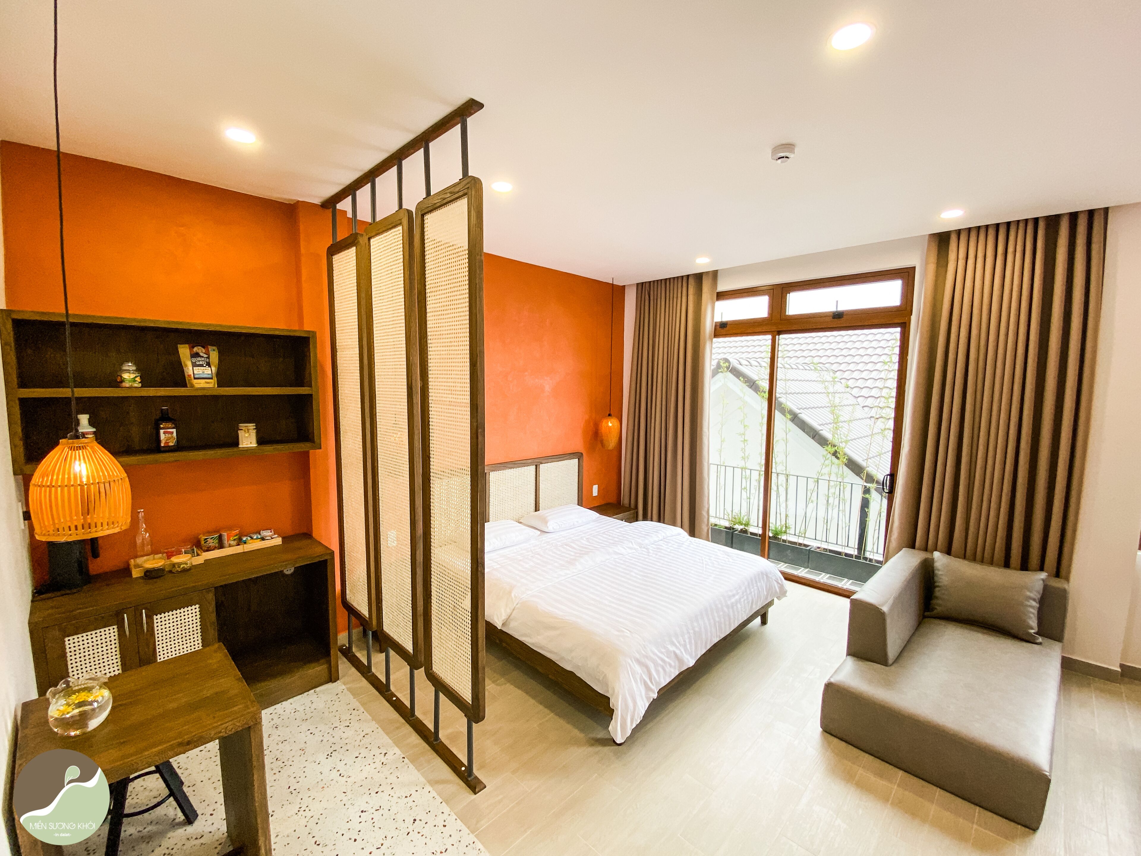 A hotel room with a bed, a sofa, a desk with a lamp, shelves with items, and a window with curtains.