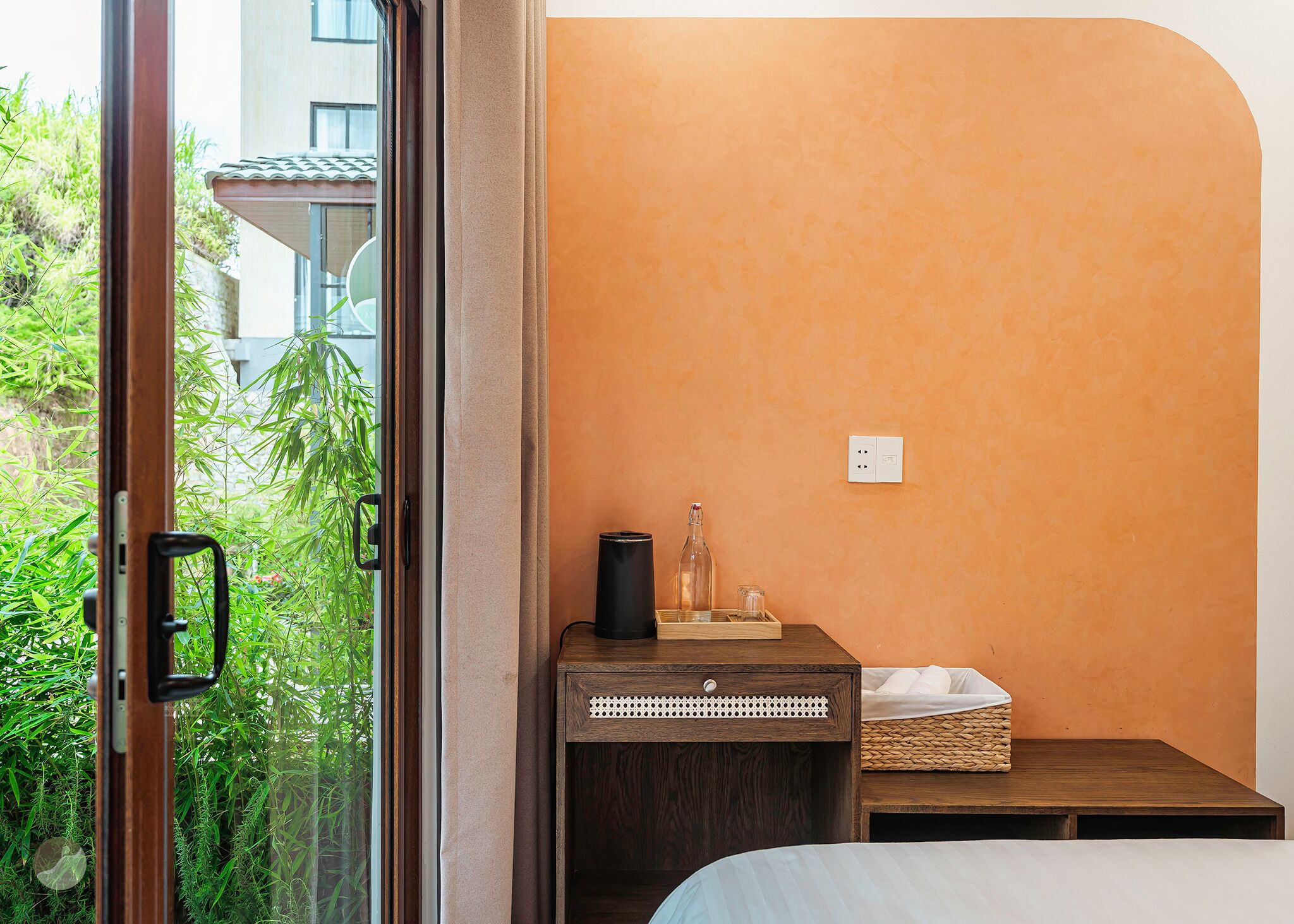 Una habitación con cama, mesita de noche, ventana con vistas a zonas verdes y en la pared, un enchufe.