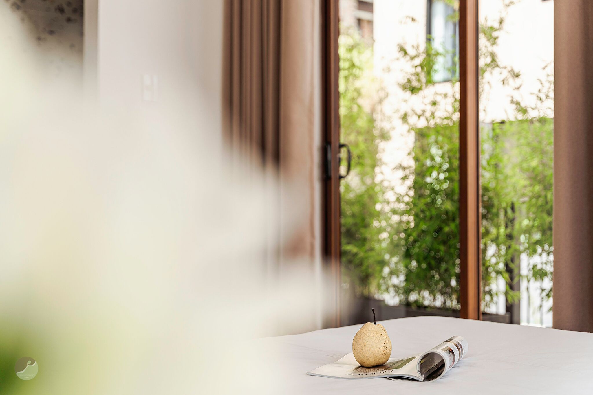 A table with a pear and a rolled-up newspaper in a room with large windows and a view of greenery outside.