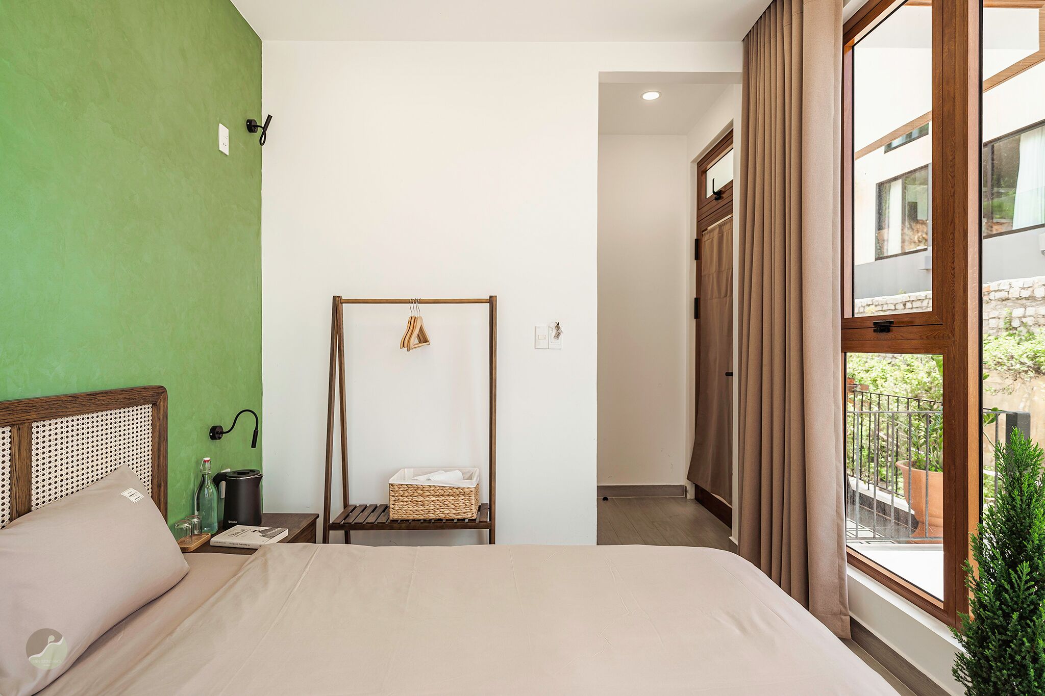 A hotel room with a bed, a bedside table, a green wall, a wooden headboard, a window with curtains, and a view of the outside.