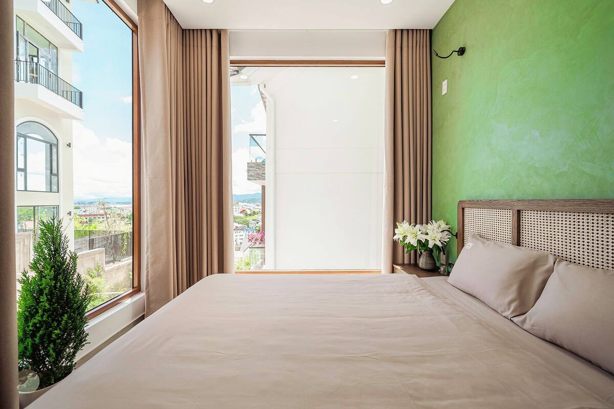A hotel room with a large bed, a view of the city through a balcony, and a green wall.