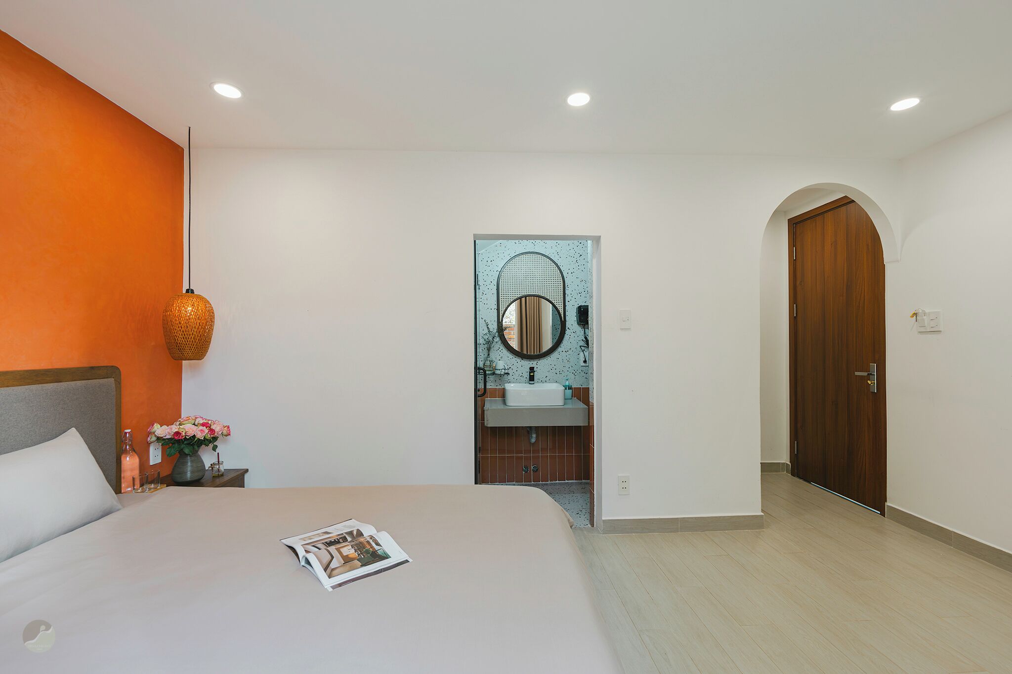 A hotel room with a bed, a nightstand with a vase of flowers, a bathroom with a mirror and sink, and a wooden door.