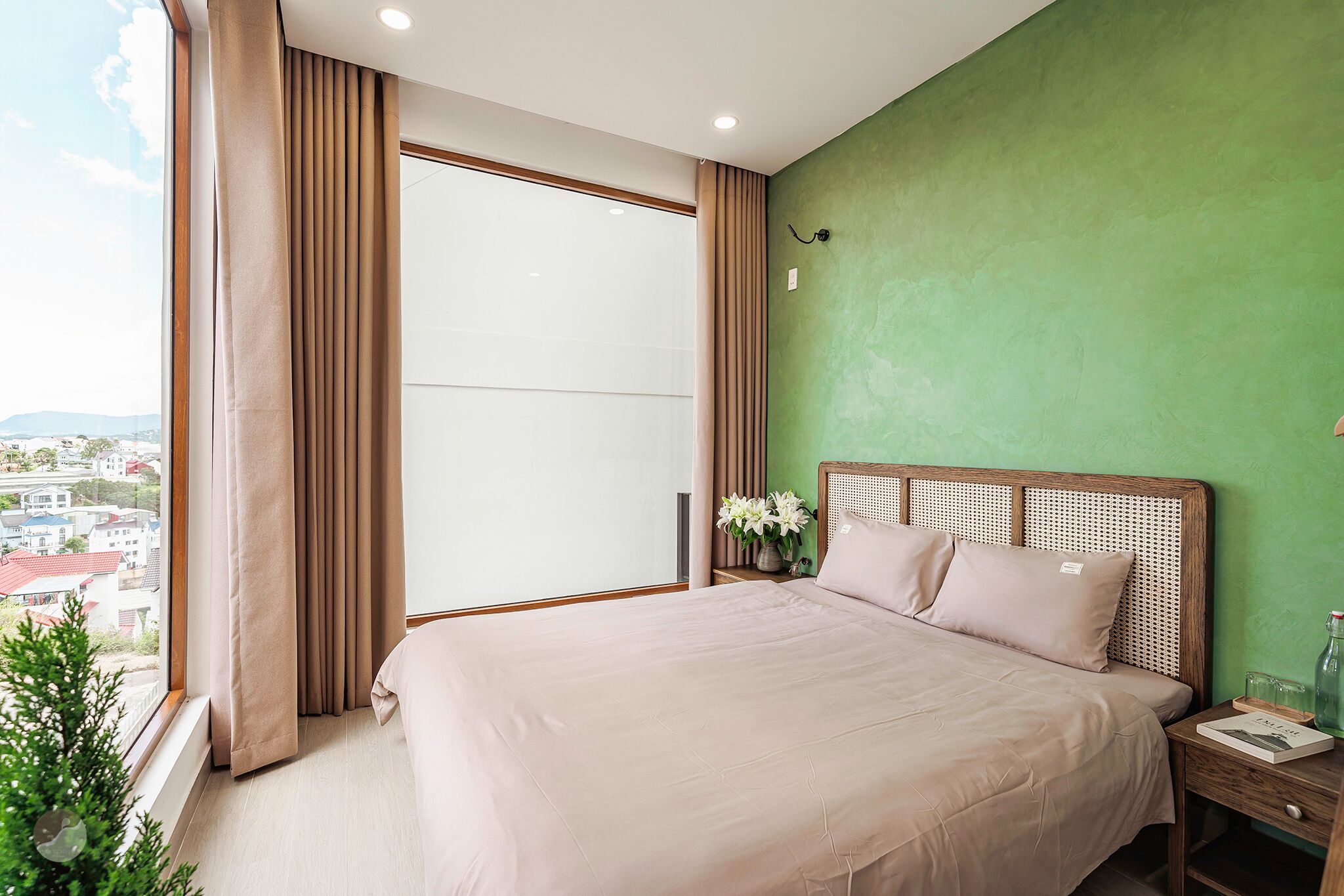 A bedroom with a large bed, a nightstand, a vase with flowers, and a view of buildings through a window.