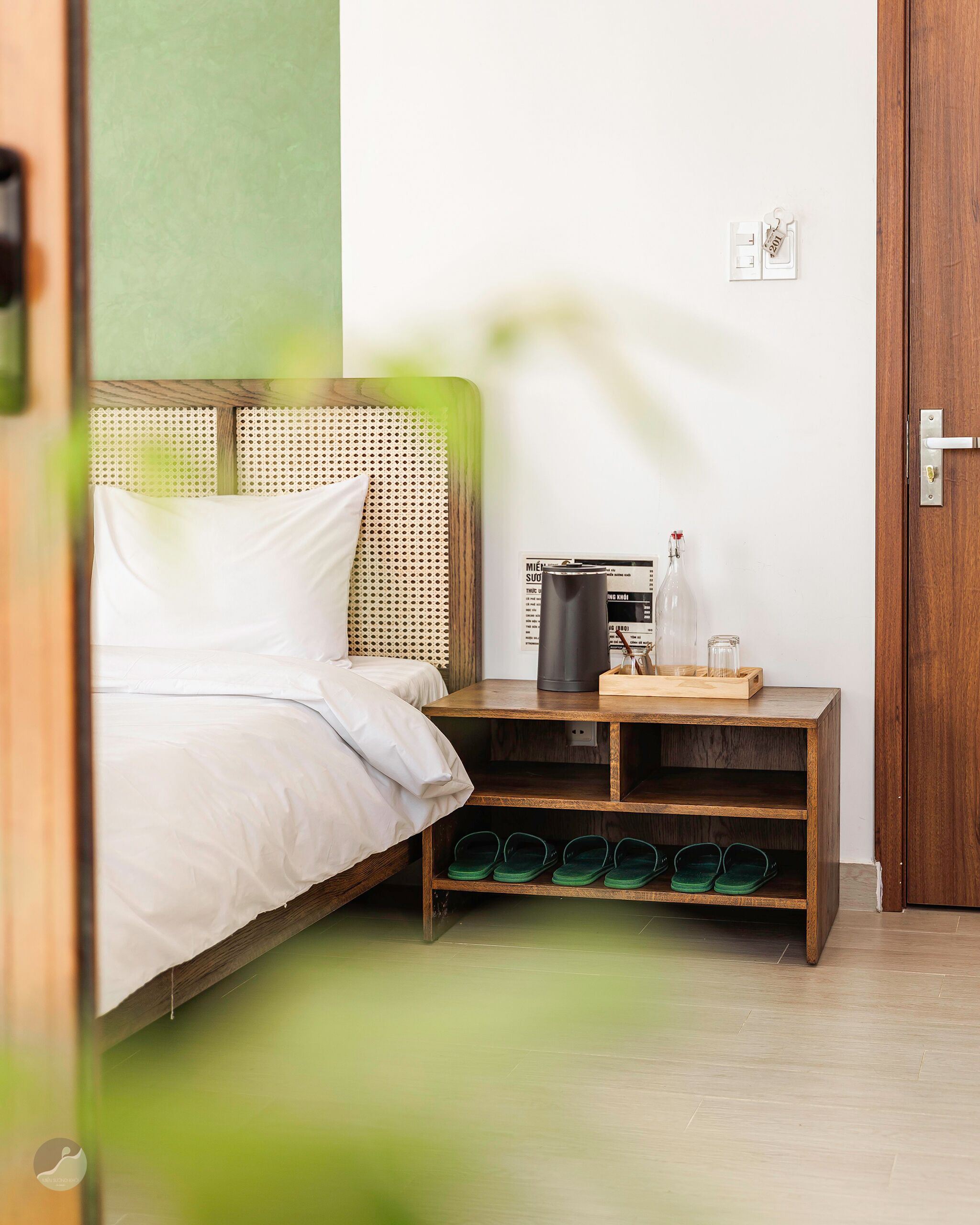 A room with a bed, a nightstand with a kettle, a bottle, and slippers.