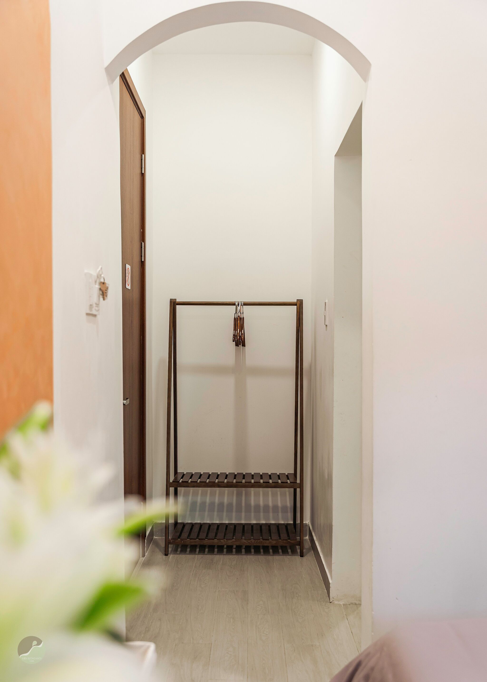 A narrow hallway with a metal bed frame at the end, a door on the left, and a wall-mounted light fixture.