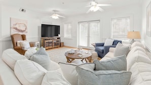 A spacious living room with a large white sofa, a blue armchair, a wooden coffee table, and a television. - Brass Rail 106 | Enjoy an Easy Stroll to the Beach or Pool! (Tybee Island)