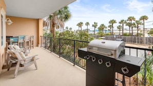 A balcony with a grill, chairs, and a view of palm trees and buildings. - Brass Rail 106 | Enjoy an Easy Stroll to the Beach or Pool! (Tybee Island)