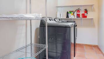 Un cuarto de lavandería con dos lavadoras apiladas, estantes metálicos para secar ropa y repisas con útiles de limpieza.