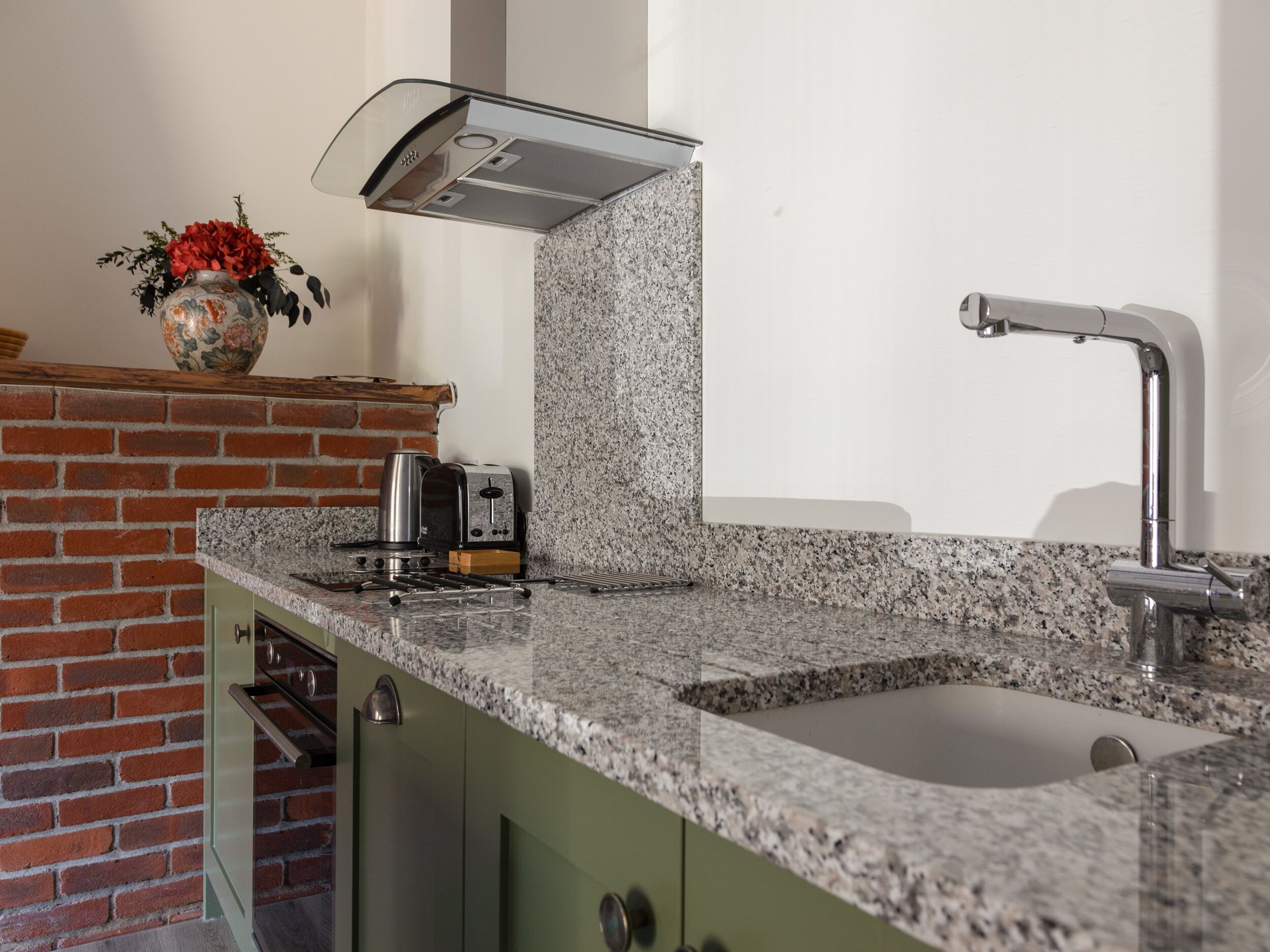 Een keuken met een granieten aanrecht, een roestvrijstalen afzuigkap, een gootsteen met een moderne kraan en een bakstenen muur.