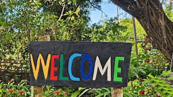 Une enseigne en bois sur laquelle est inscrit le mot « BIENVENUE » en lettres colorées, le tout sur fond de verdure luxuriante et avec un arbre en arrière-plan.