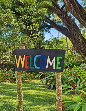 Un letrero de madera con la palabra 'WELCOME' en letras coloridas, frente a un fondo de vegetación exuberante y un árbol.