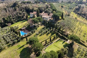 Eine Vogelperspektive zeigt eine Villa mit Tennisplatz, umgeben von üppiger Vegetation und hoch aufragenden Zypressen.