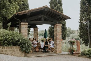 Area esterna coperta con pilastri in pietra e tetto in legno, immersa nel verde e accessibile tramite un vialetto ghiaioso.