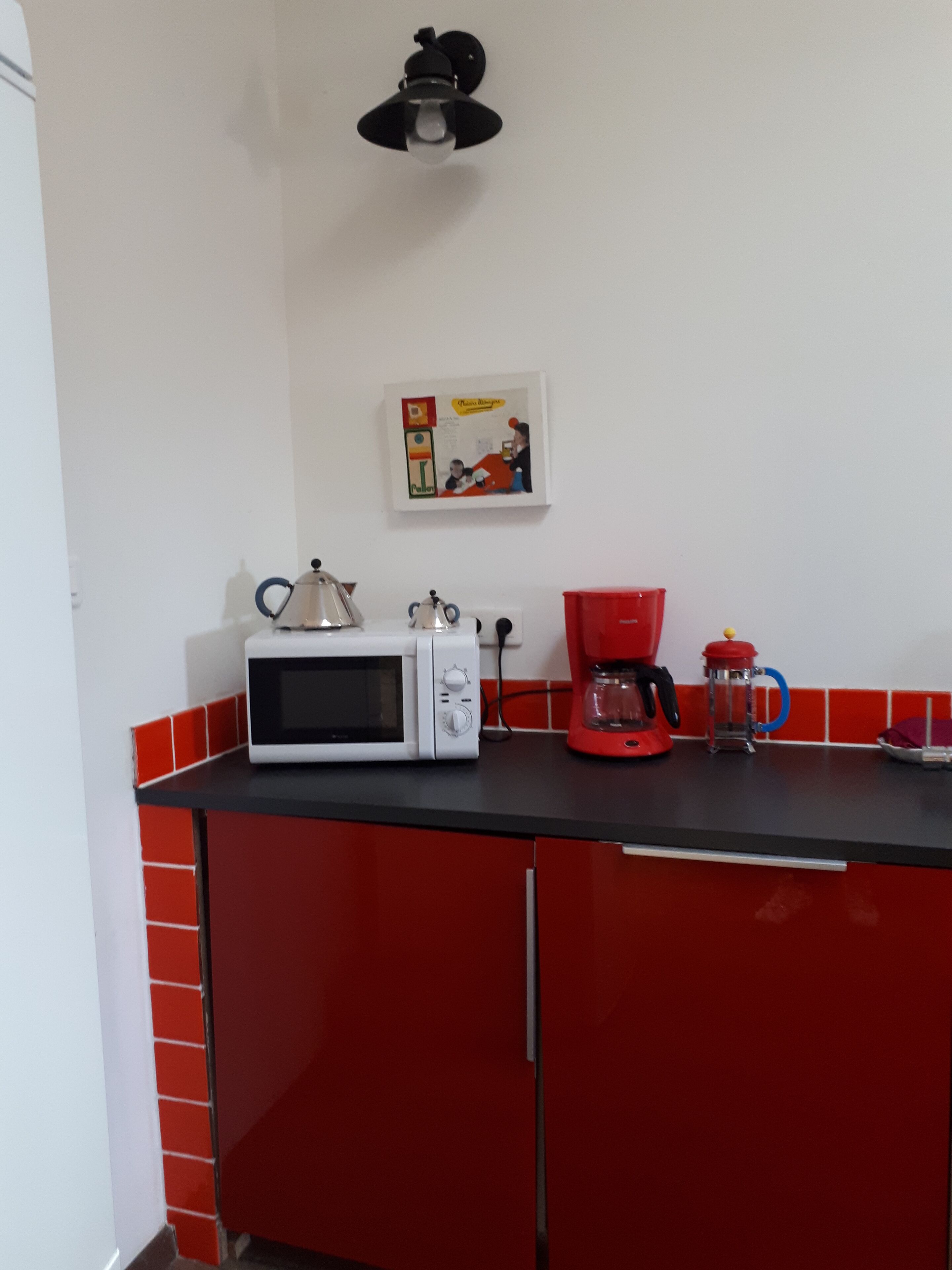 Una cocina con microondas, cafetera y hervidor en la encimera, y un cuadro en la pared.