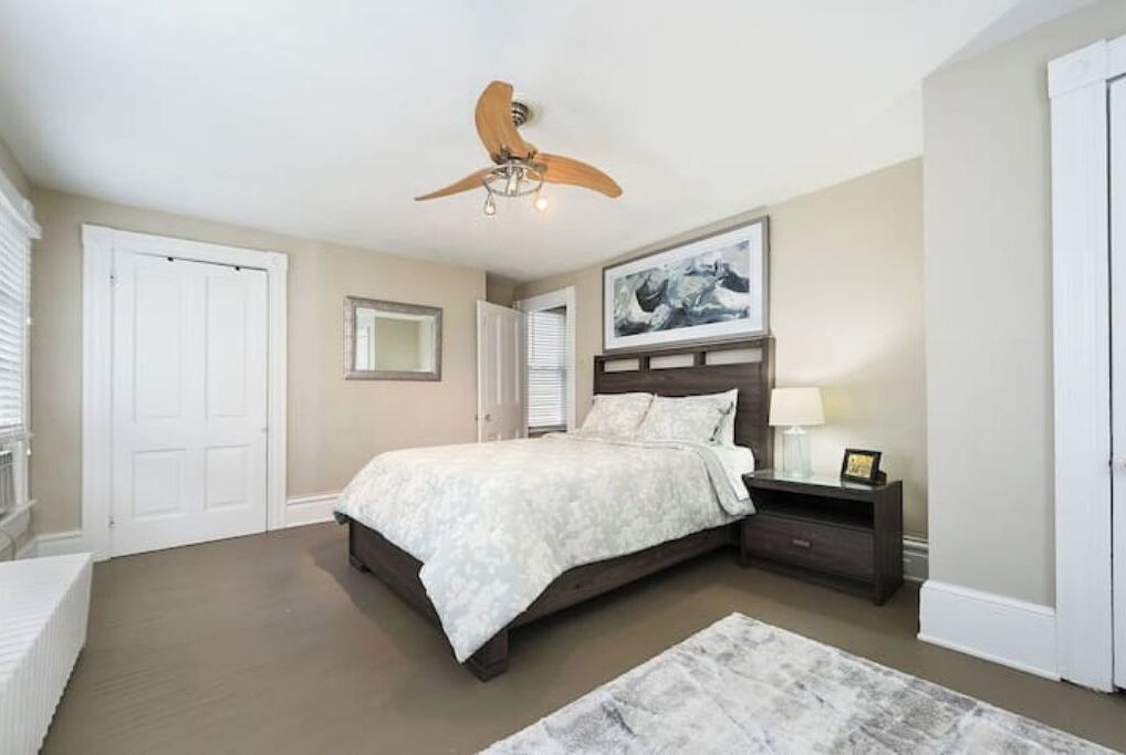 A bedroom with a bed, a nightstand, a ceiling fan, a framed picture on the wall, and a door.