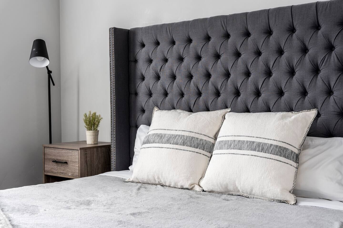A bedroom with a tufted headboard, two pillows with striped patterns, a bedside table with a lamp, and a small potted plant.