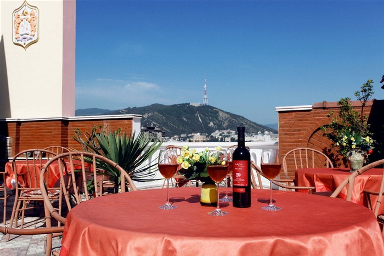 An outdoor dining area with a table set for two, a bottle of wine, and glasses on a red tablecloth. A mountain with a tower is visible in the background.