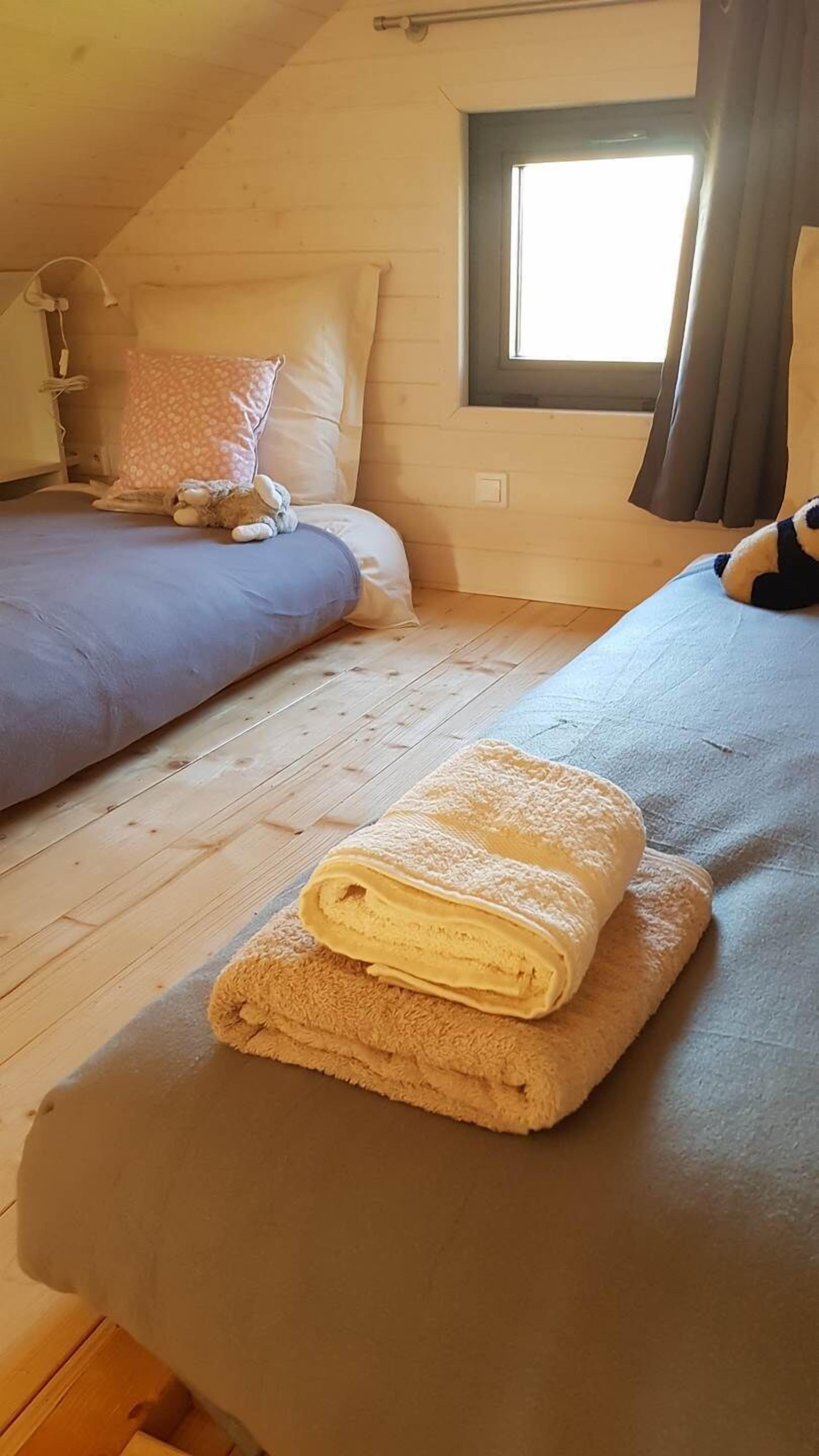 A cozy room with a wooden ceiling, a window with curtains, a bed with a rolled-up towel, and a bedside table.
