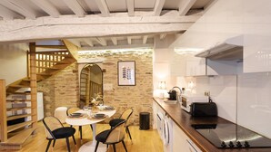 A modern kitchen with a wooden staircase, a dining area with a round table and chairs, and a stone wall. - L'Honoré - Appt T2 en duplex - centre ville (Rennes)
