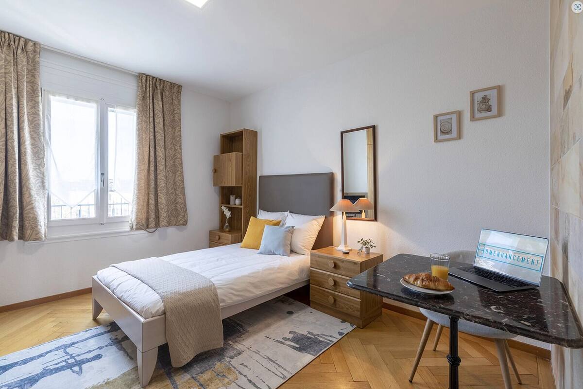 Une chambre d’hôtel avec un lit, une table de chevet, une lampe, un ordinateur portable et une petite table sur laquelle se trouvent un croissant et un jus de fruits.