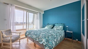 A bedroom with a large bed, a chair, a bedside table, and a view through a window. - Duplex Etoile de mer- T4 Vue mer (Courseulles-sur-Mer)