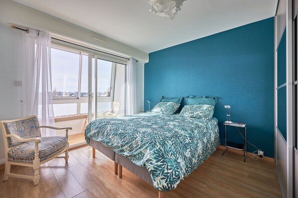 A bedroom with a large bed, a chair, a bedside table, and a view through a window.