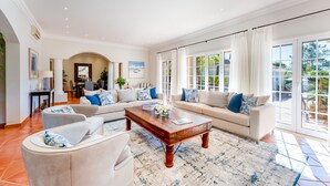 A spacious living room with a large sofa set, a coffee table, and a view of the outdoors through large windows. - Villa Breena - Quinta do Mar (Quinta do Lago)