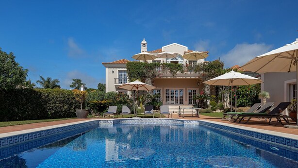 A swimming pool with lounge chairs and umbrellas, surrounded by lush greenery and a large building with a balcony. - Villa Breena - Quinta do Mar (Quinta do Lago)