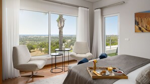Une chambre d’hôtel moderne, dotée d’un grand lit, de deux fauteuils, d’une petite table ronde et offrant une vue sur la campagne grâce à de grandes fenêtres.