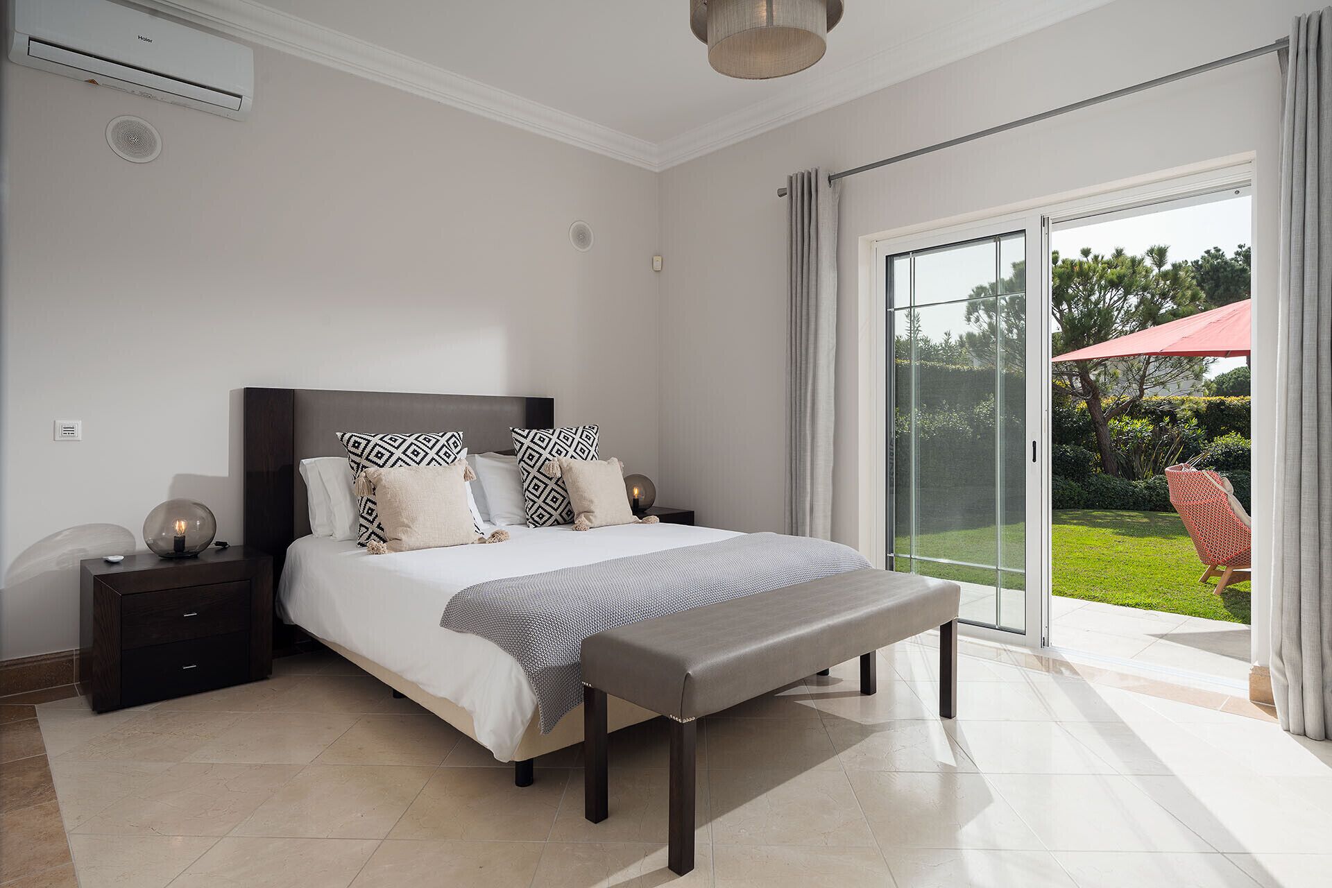 A bedroom with a large bed, a bench, a nightstand, and a view of a garden through a sliding glass door.