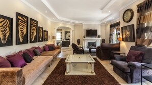 A spacious living room with a large sofa, a coffee table, and a fireplace. There are framed artworks on the walls and a television mounted above the fireplace.