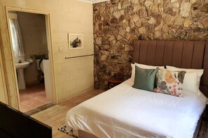 A bedroom with a stone wall, a bed with pillows, a nightstand, and a bathroom visible through an open door.