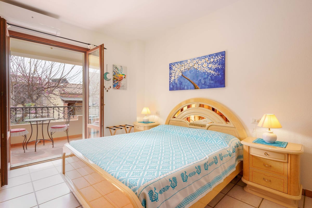 Une chambre à coucher comprenant un lit, une table de chevet, une lampe, un tableau et un balcon avec une table et des chaises.