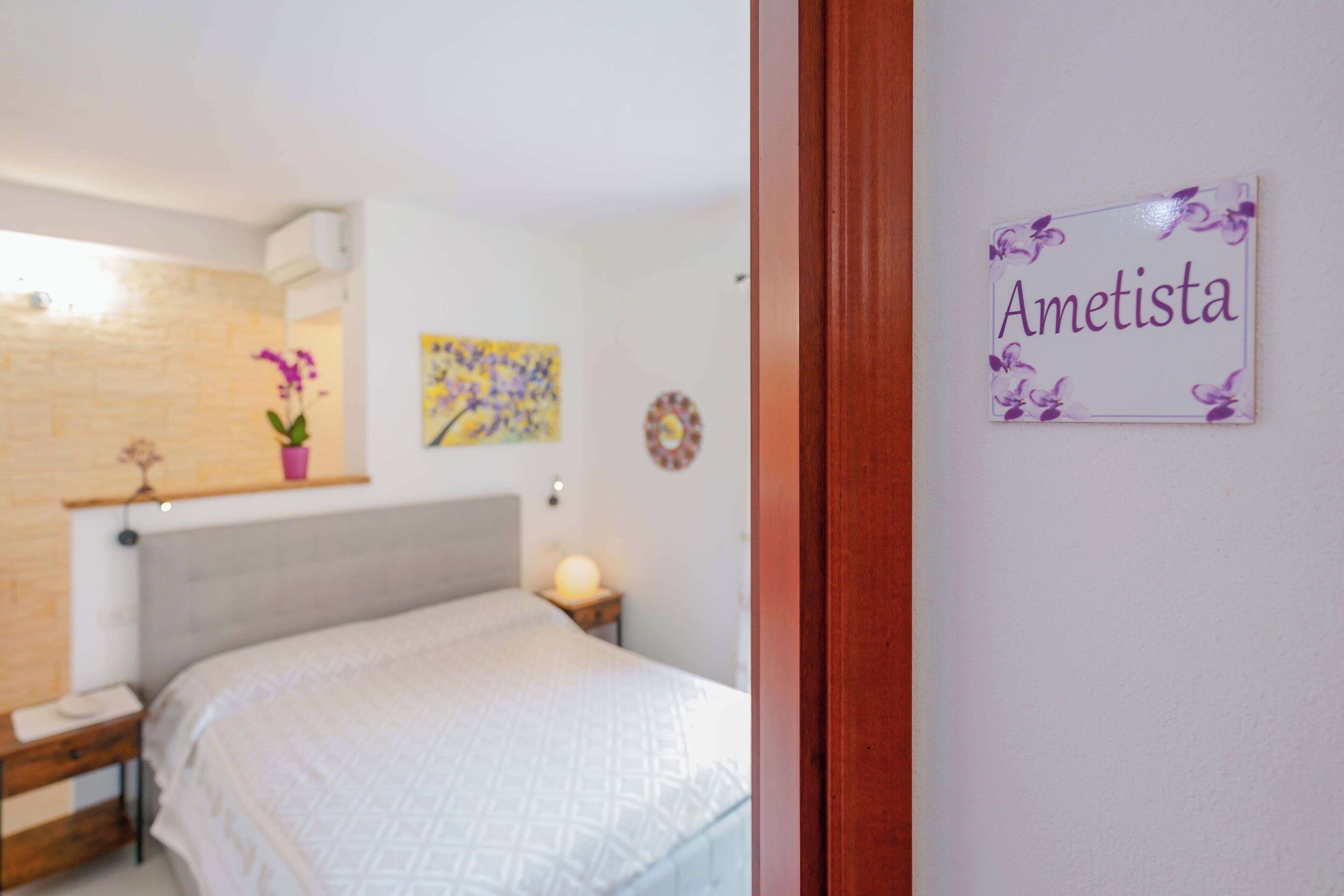 Un dormitorio con cama, mesita de noche, estante de pared con planta en maceta, reloj de pared y placa con el nombre 'Ametista'.