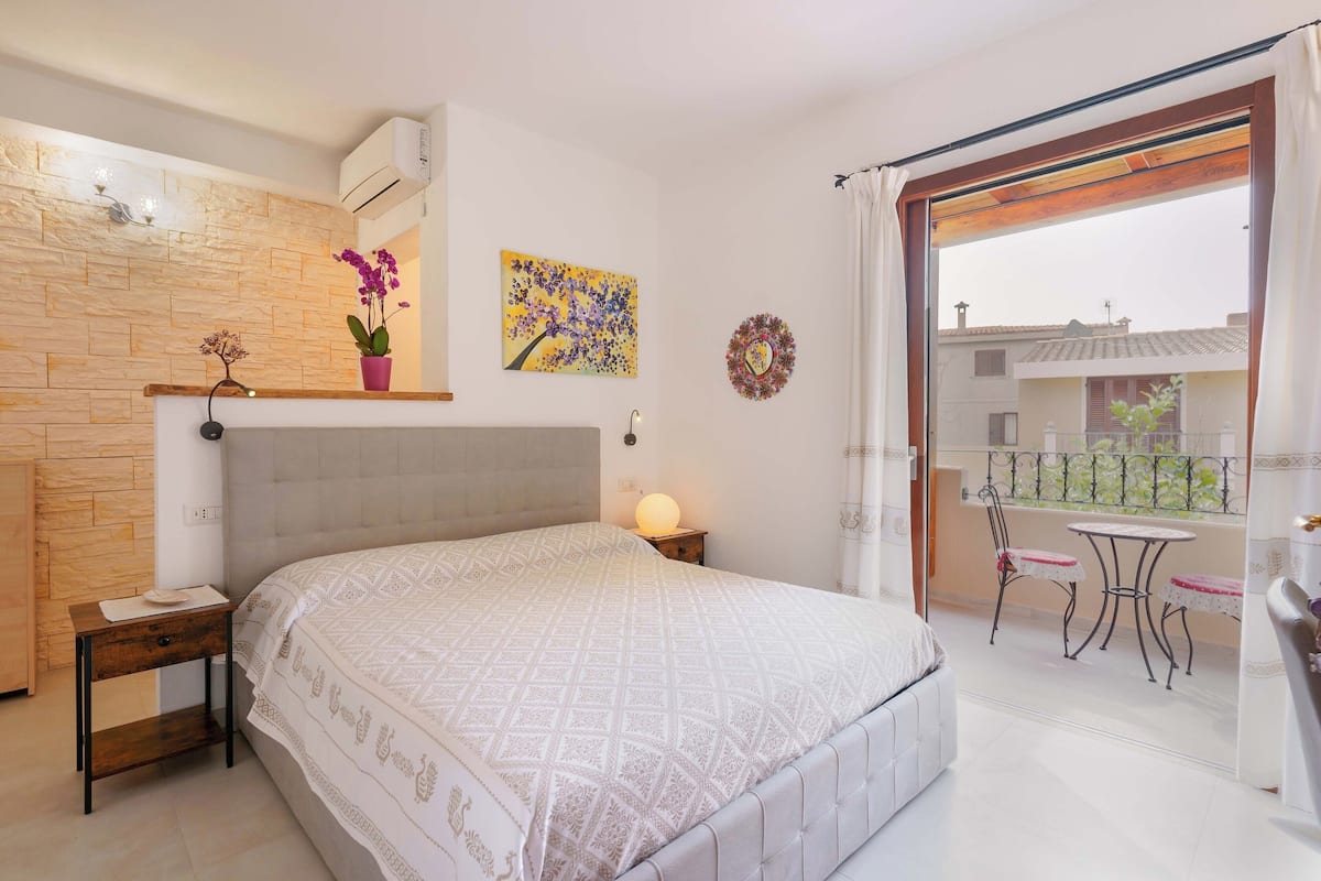 Une chambre comprenant un lit, une table de chevet, un climatiseur mural, une orchidée en pot, un élément décoratif mural et un balcon avec une table et des chaises.