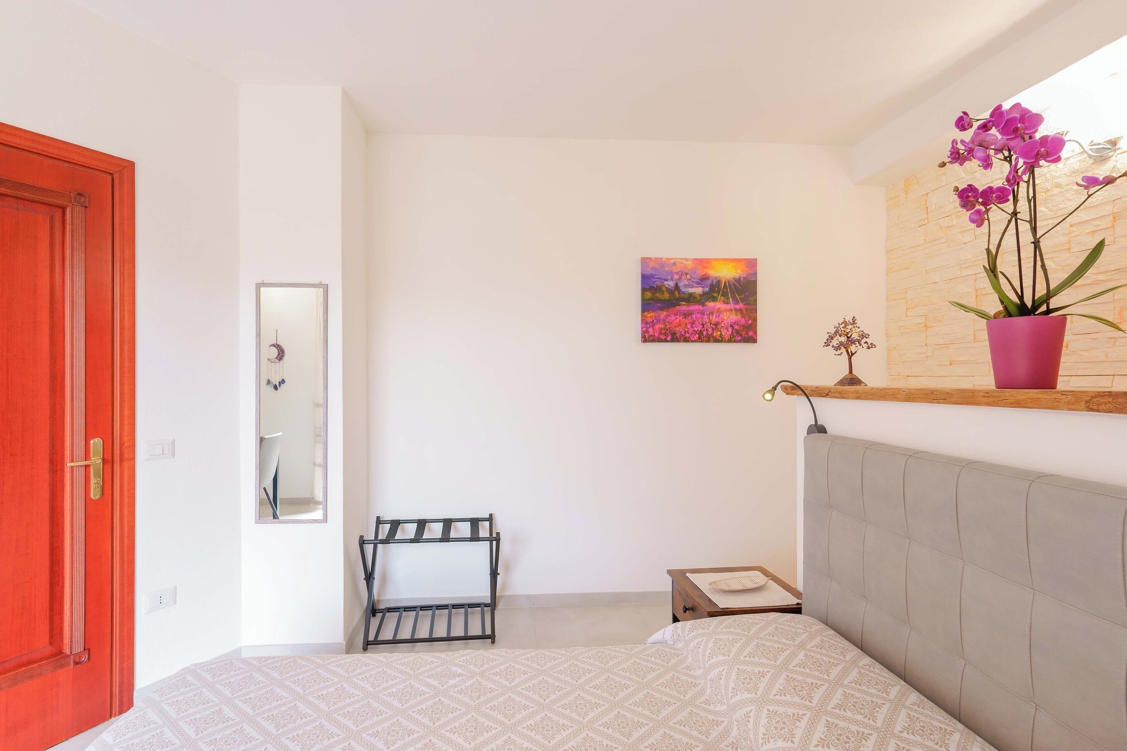 Un dormitorio con una cama, una puerta roja, un espejo, un cuadro y una orquídea en maceta.