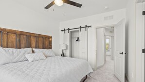 A bedroom with a bed, a ceiling fan, a wooden headboard, a white door, and a hallway leading to another room.