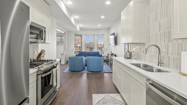 A modern kitchen with stainless steel appliances, a dining area with a blue sofa, and a view of the city through large windows.