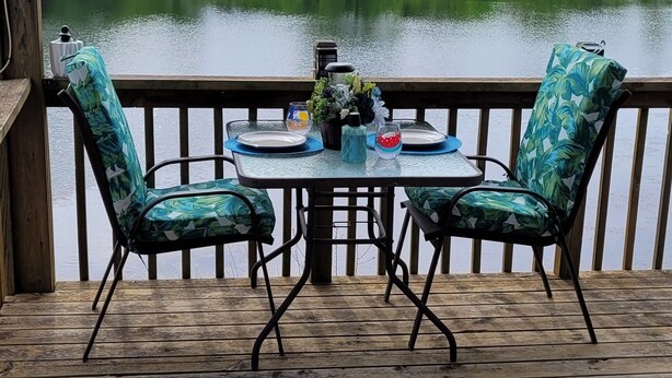 Une terrasse en bois avec deux chaises et une table, donnant sur un lac bordé d’arbres.