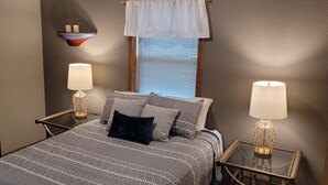 Une chambre à coucher avec un lit, deux tables de chevet avec des lampes, une petite table, une étagère murale avec des bougies et une fenêtre avec des stores.